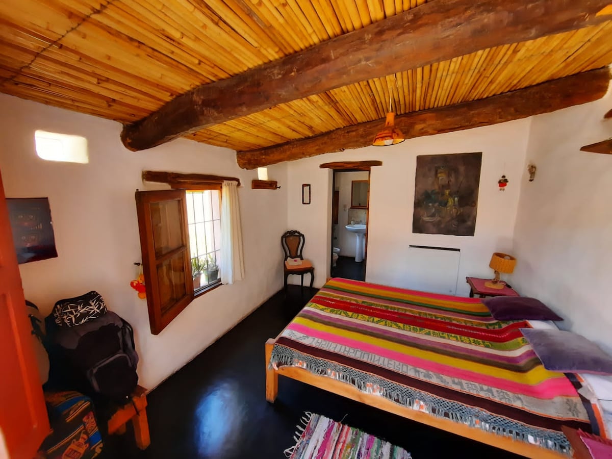 A bedroom features a spacious bed topped with colorful blankets. Exposed wooden beams and a thatched ceiling add character. Natural light filters through windows, illuminating the neutral walls. A chair and a small table complement the cozy space, creating a welcoming environment.