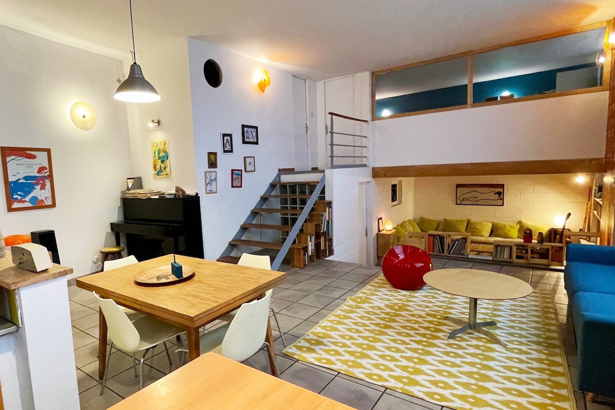 A spacious loft interior features a large dining table surrounded by white chairs and a cozy seating area with a blue couch. A patterned rug lies on the tiled floor, while stairs lead to a mezzanine. A piano and shelves add to the welcoming ambiance.