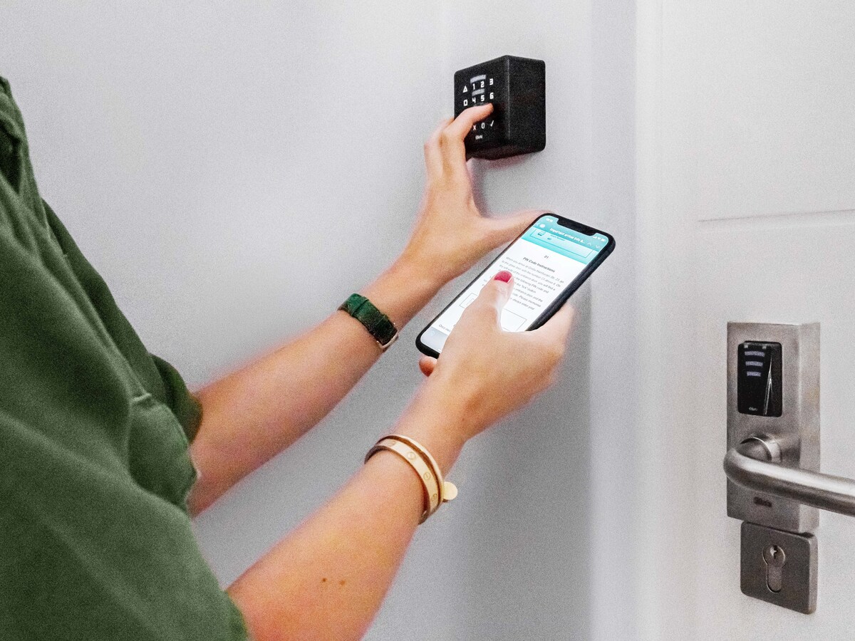 A hand is seen entering a digital PIN code on a black keypad mounted on the wall. A smartphone is held in the other hand, displaying an app with instructions. The scene illustrates a contact-free self-check-in process.