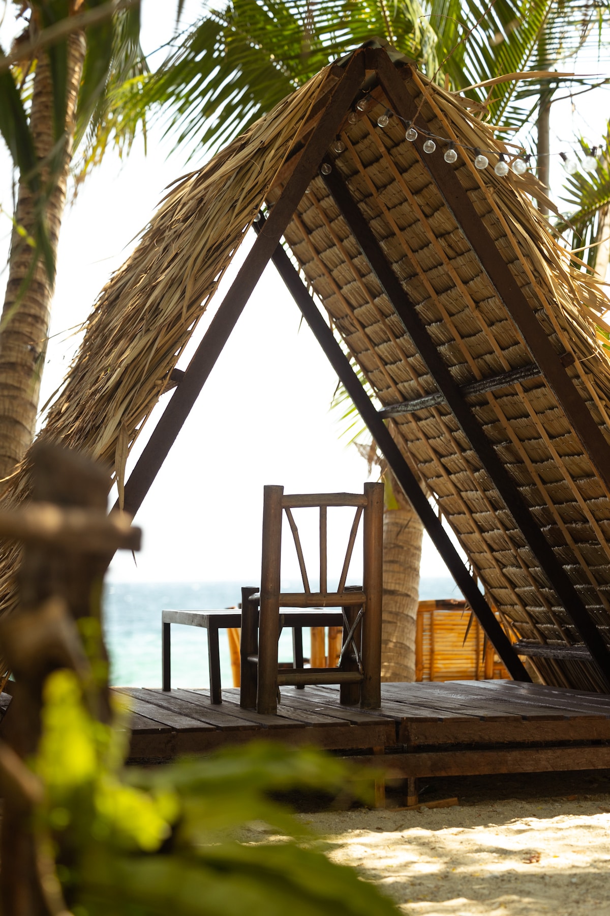 A sheltered seating area is framed by palm trees and features a rustic wooden chair under a thatched roof. Sunlight filters through the structure, highlighting the sandy surroundings and the serene sea view in the background.