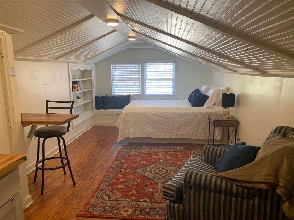 A well-lit attic space features a queen bed with white bedding. Windows provide natural light, and a small table with a bar stool is positioned nearby. A striped chair and a decorative area rug add comfort to the room, showcasing a blend of functionality and style.
