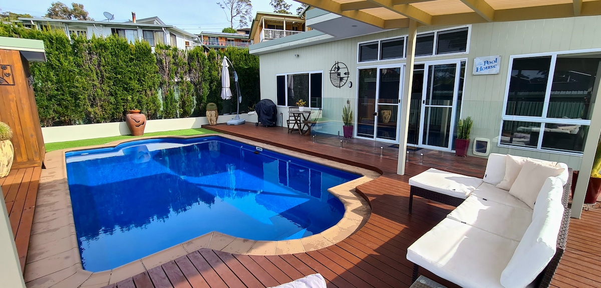 A private outdoor area features a spacious pool surrounded by wooden decking. A lounge area includes a cushioned seating arrangement, and a dining table is set against the backdrop of lush greenery. Large glass doors provide access to the interior space.