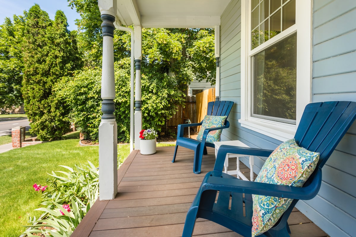 A charming front porch features two blue Adirondack chairs, each adorned with colorful cushions. Potted plants are placed nearby, and a lush green lawn and tall shrubs create a serene outdoor setting. The porch is framed by white columns, inviting relaxation.
