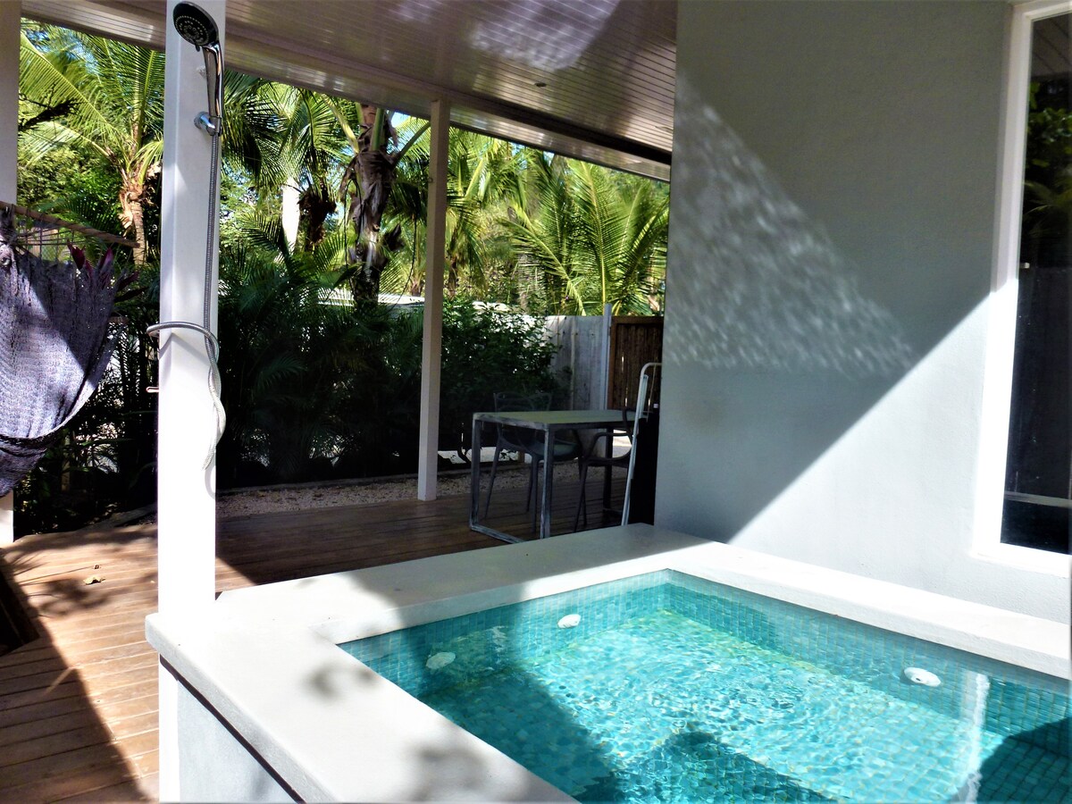 A private pool is featured adjacent to a spacious wooden deck, surrounded by lush tropical vegetation. A dining area with a table is visible under the covered patio, offering protection from the sun.