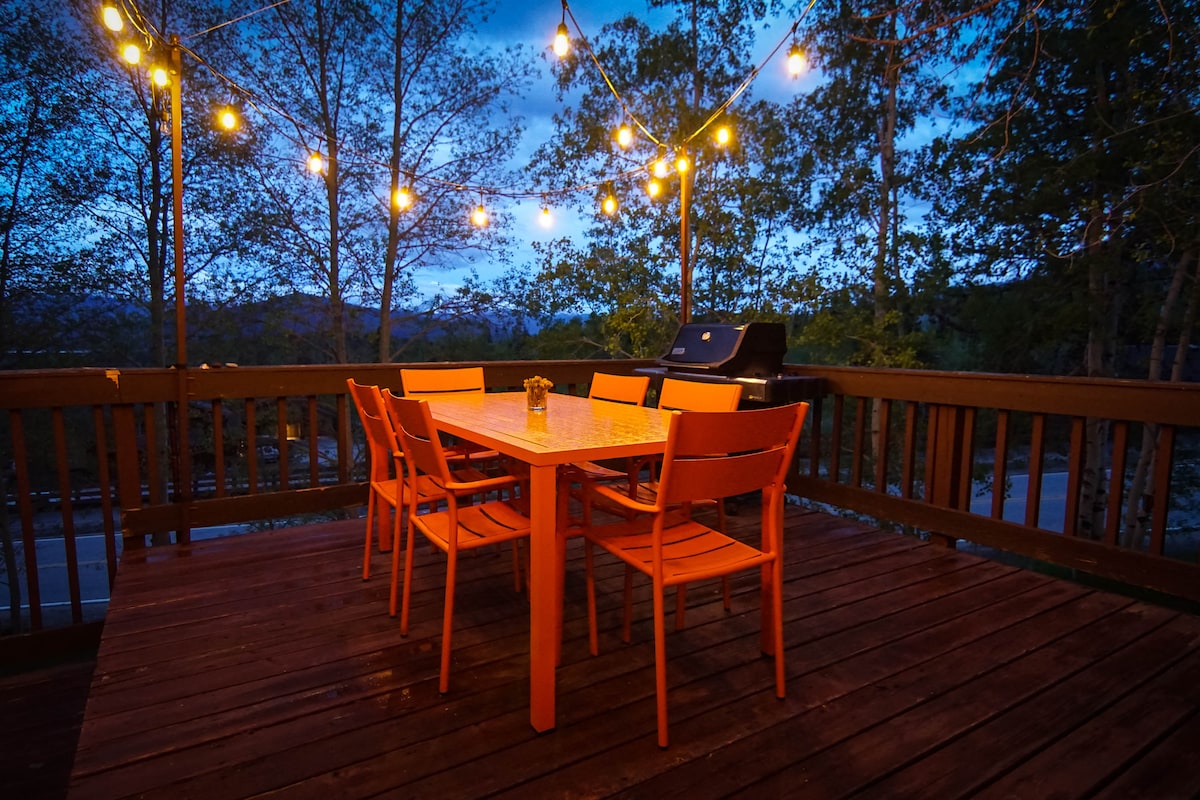 A spacious deck features a large table surrounded by six orange chairs, providing a welcoming dining area. String lights overhead create a warm ambiance, while a grill stands ready for outdoor cooking. Lush trees frame the setting, enhanced by the evening sky.