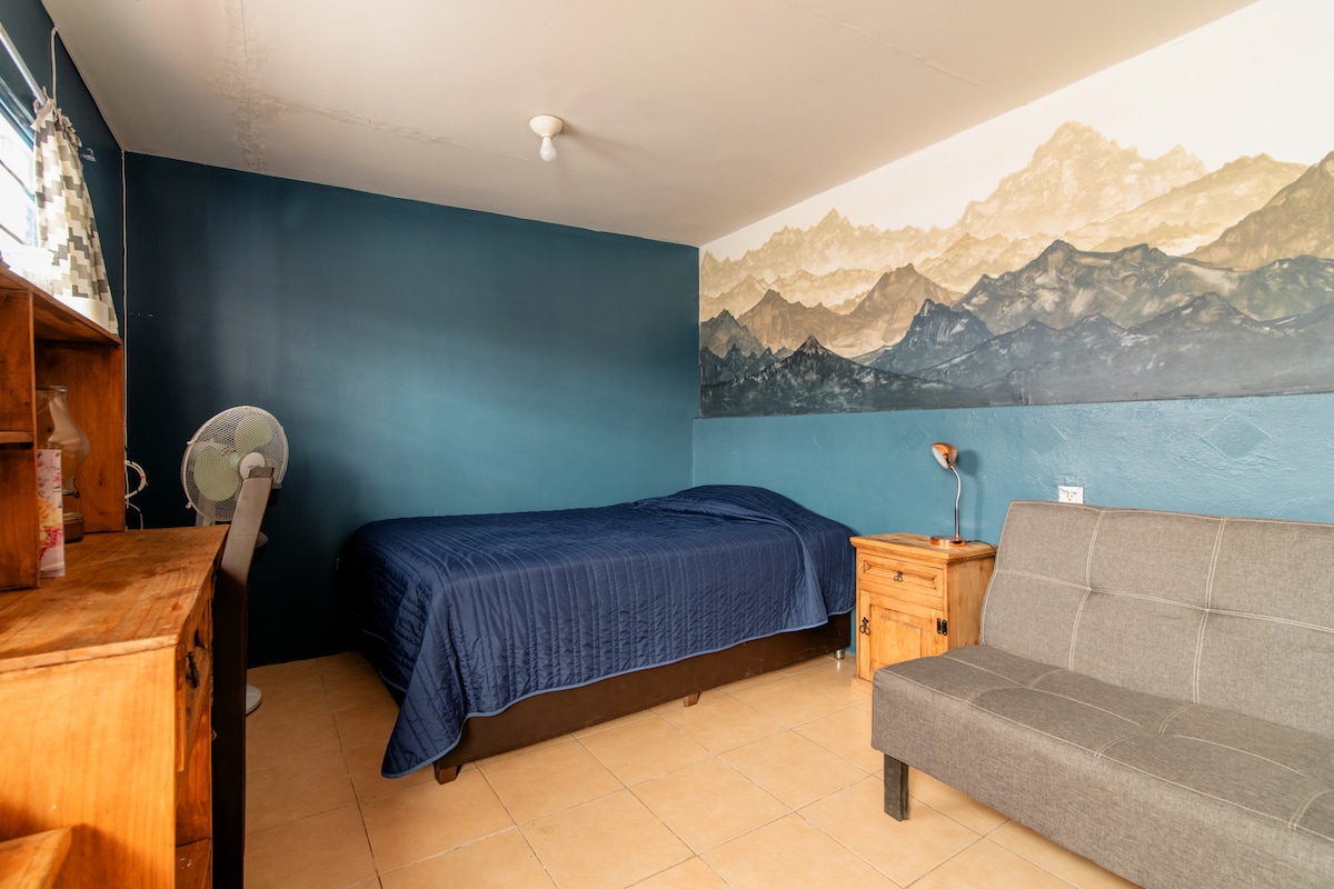 A sleeping area features a spacious bed covered with a blue blanket, complemented by a mural of mountain landscapes on one wall. A wooden desk and nightstand are present, alongside a gray sofa and a fan, creating a functional and inviting space.