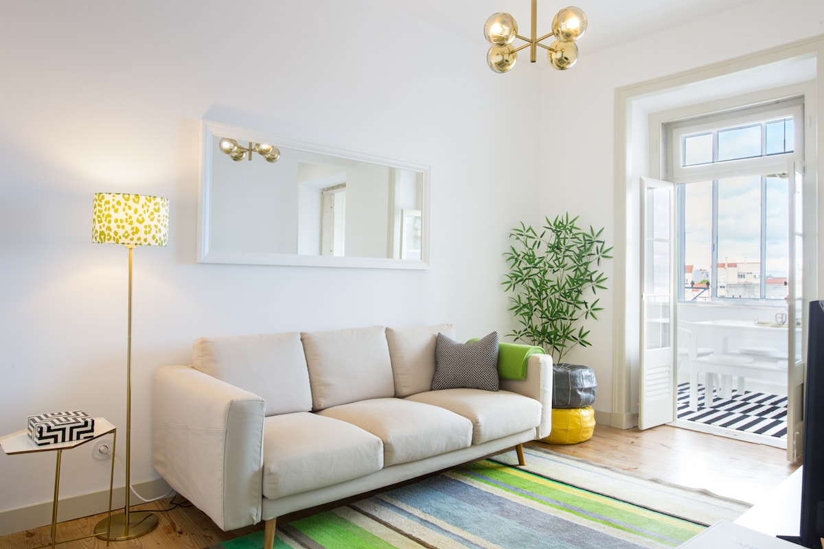 A cozy living room features a light-coloured sofa paired with a grey pillow. A stylish floor lamp with a patterned shade adds a pop of colour. A striped area rug lies beneath a coffee table. Bright natural light enters through large windows that open to a balcony.