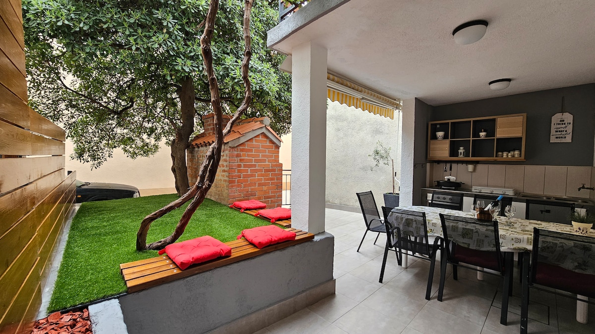 An outdoor terrace is depicted, featuring a dining area with a table and chairs under a covered area. Lush greenery is present, alongside low seating cushions on wooden platforms. A brick structure lies in the background, contributing to a relaxed ambiance.
