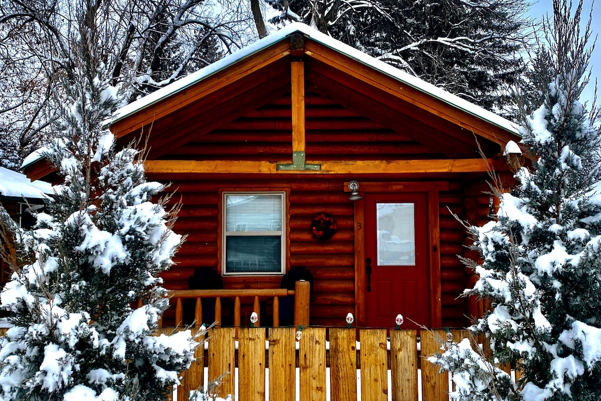A charming log cabin is surrounded by snow-covered evergreens, showcasing a wooden porch and a red door. The cabin's rustic design is highlighted by its wood siding and a festive wreath on the door, creating a warm welcome amidst the winter landscape.