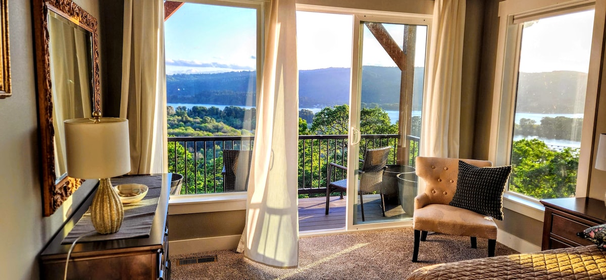 A bright bedroom features large sliding glass doors that open to a private balcony. Sunlight filters through sheer curtains, highlighting the stylish decor, including a comfortable chair and decorative lamp. The view beyond showcases lush trees and the serene waters of the Columbia River.