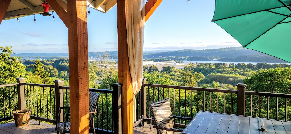 A spacious deck is framed by wooden columns, offering a panoramic view of the lush landscape below, including rivers and mountains. Outdoor seating is arranged around a table, with an umbrella providing shade. The setting is illuminated by soft outdoor lights.