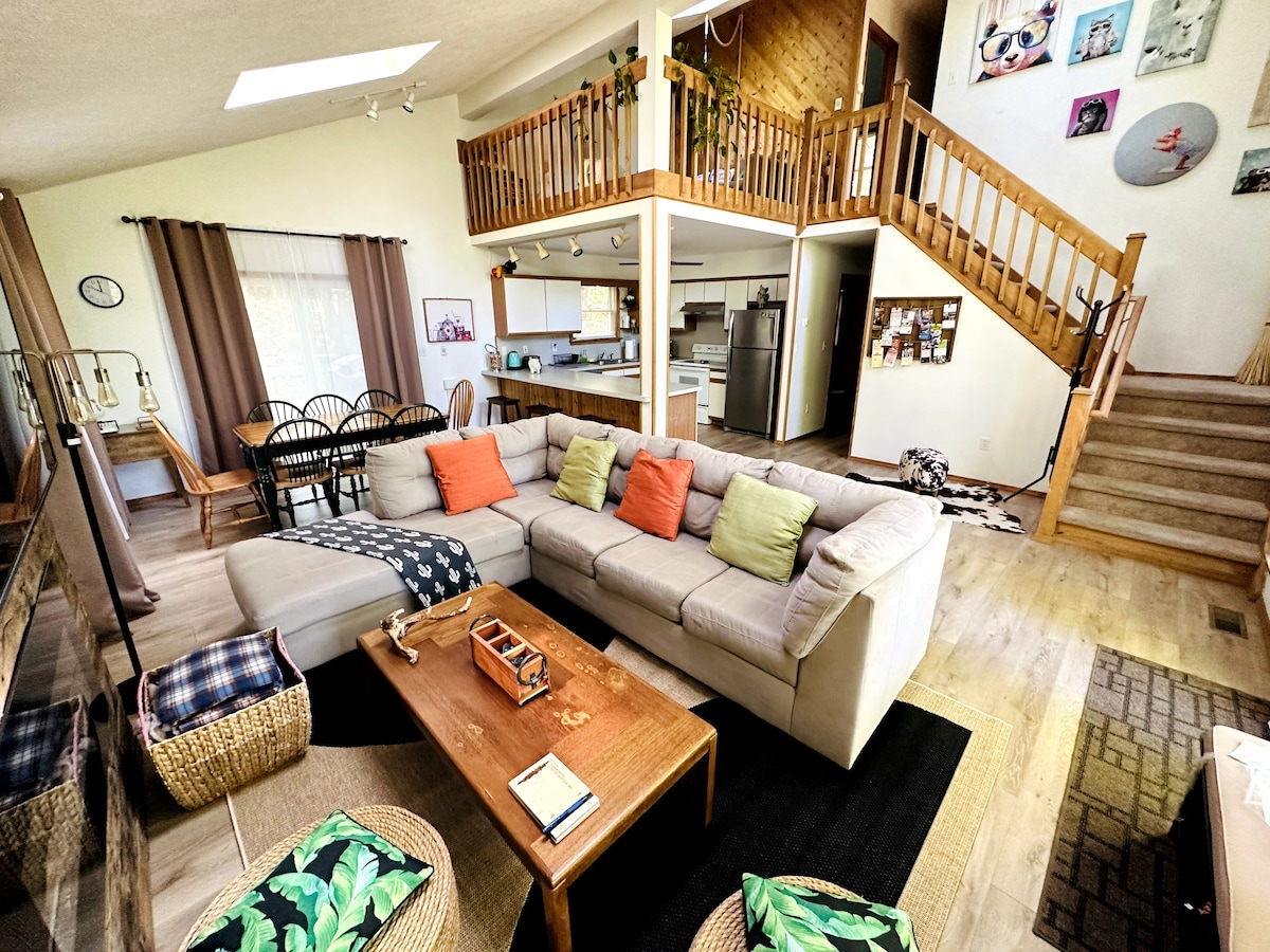 A spacious living area features a large, light-colored sectional sofa adorned with colorful cushions. A wooden coffee table sits at the center, surrounded by woven storage baskets. In the background, an open concept kitchen and dining area can be seen, with a staircase leading to the upper level.