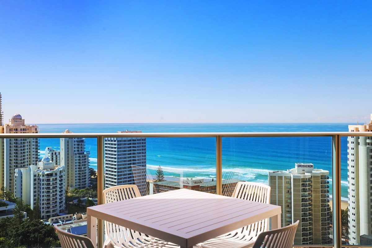 A private balcony features a white outdoor dining table and chairs, overlooking a panoramic view of the ocean and cityscape. The clear blue sky complements the serene waters, creating a tranquil setting for relaxation.