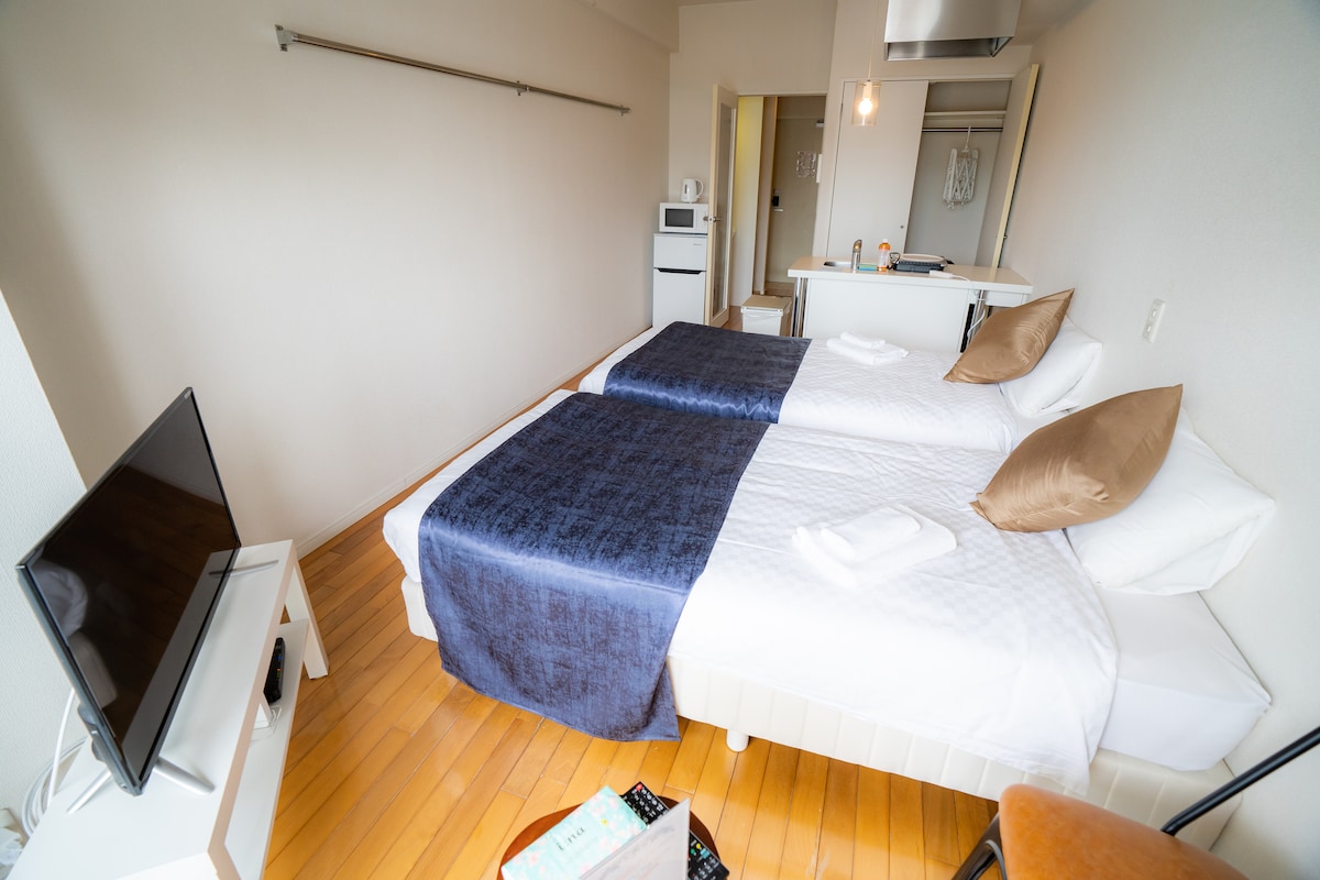 A compact studio features two spacious beds with crisp white linens and decorative brown pillows. A flat-screen TV is positioned on a white stand, and a small kitchenette is visible in the background, enhancing the functional layout.