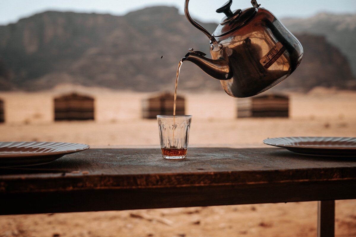 A traditional kettle is seen pouring tea into a glass, positioned on a rustic wooden table. Two plates are set on the table, with a backdrop of the desert landscape and distant seating areas visible in the background.