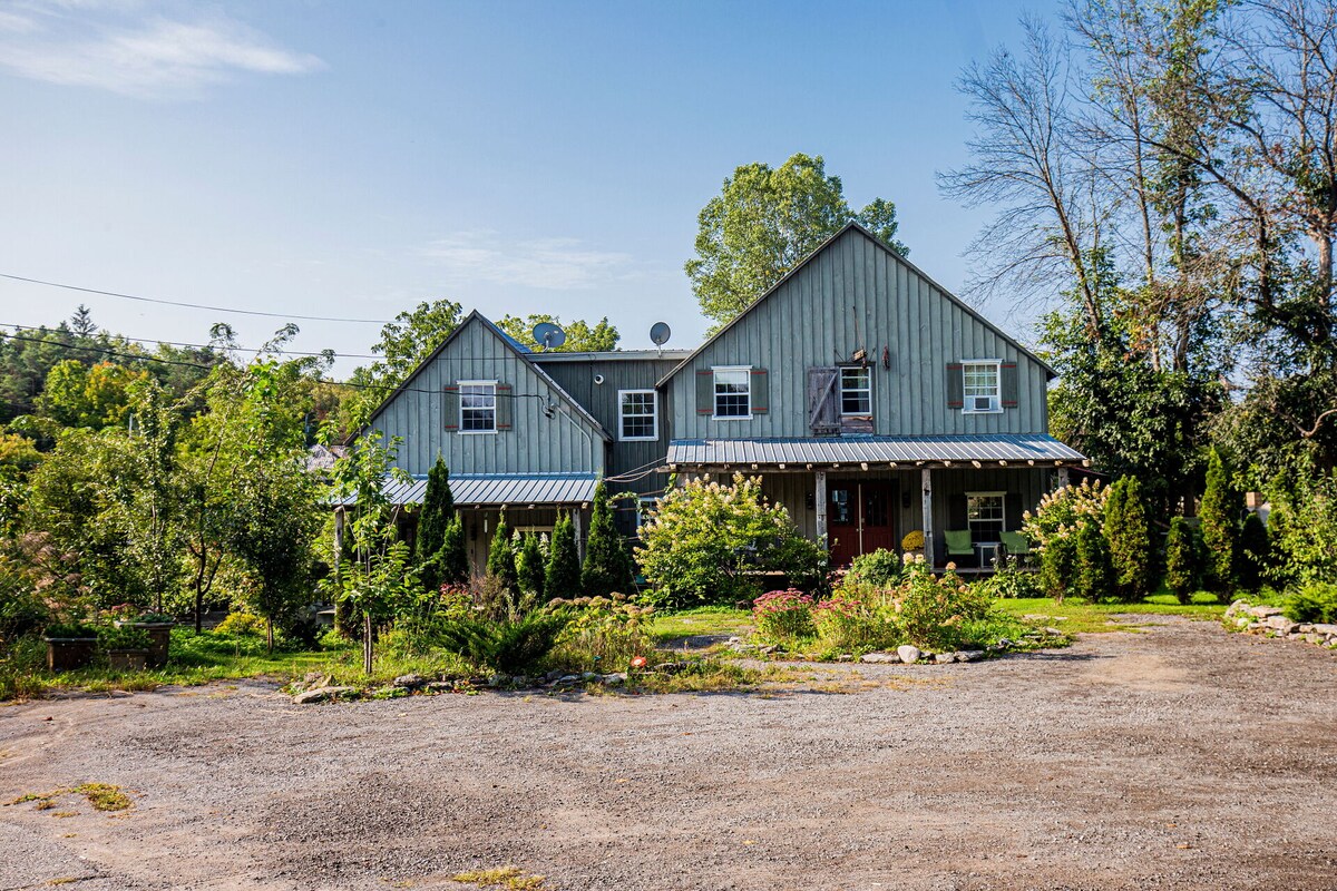 A rustic two-story cottage is framed by lush greenery, with a welcoming porch adorned by potted plants. The building features multiple windows and a metal roof, while a gravel area in front offers space for parking. The natural surroundings contribute to a serene atmosphere.