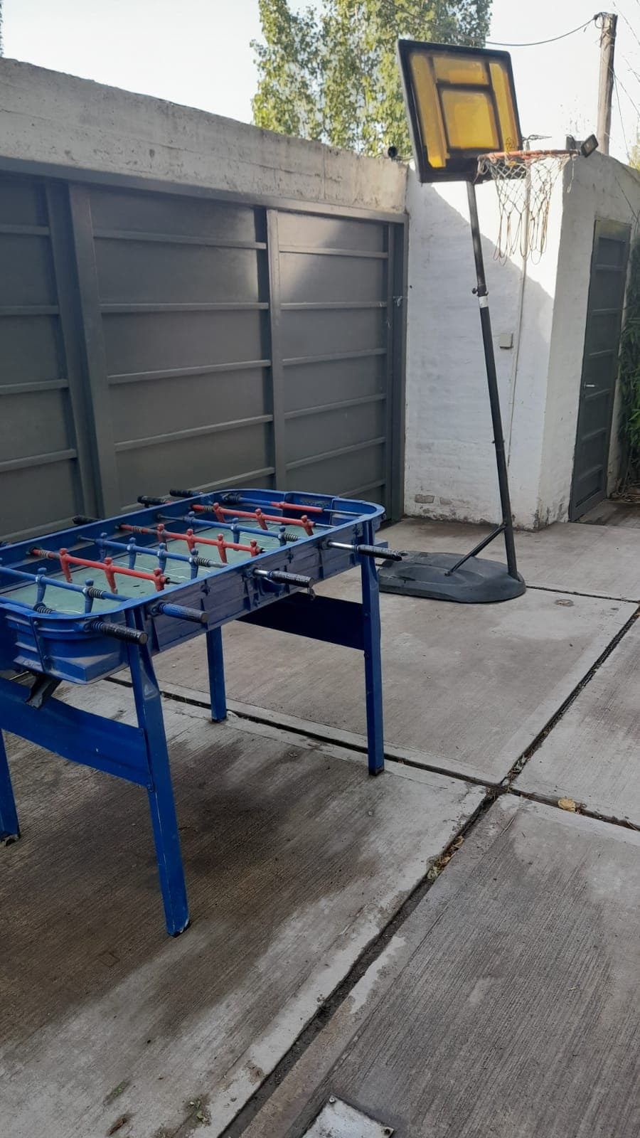 An outdoor area features a blue foosball table and a basketball hoop. The surfaces are made of concrete, and the space is enclosed by walls. Natural light illuminates the scene, creating a casual and inviting environment for outdoor activities.