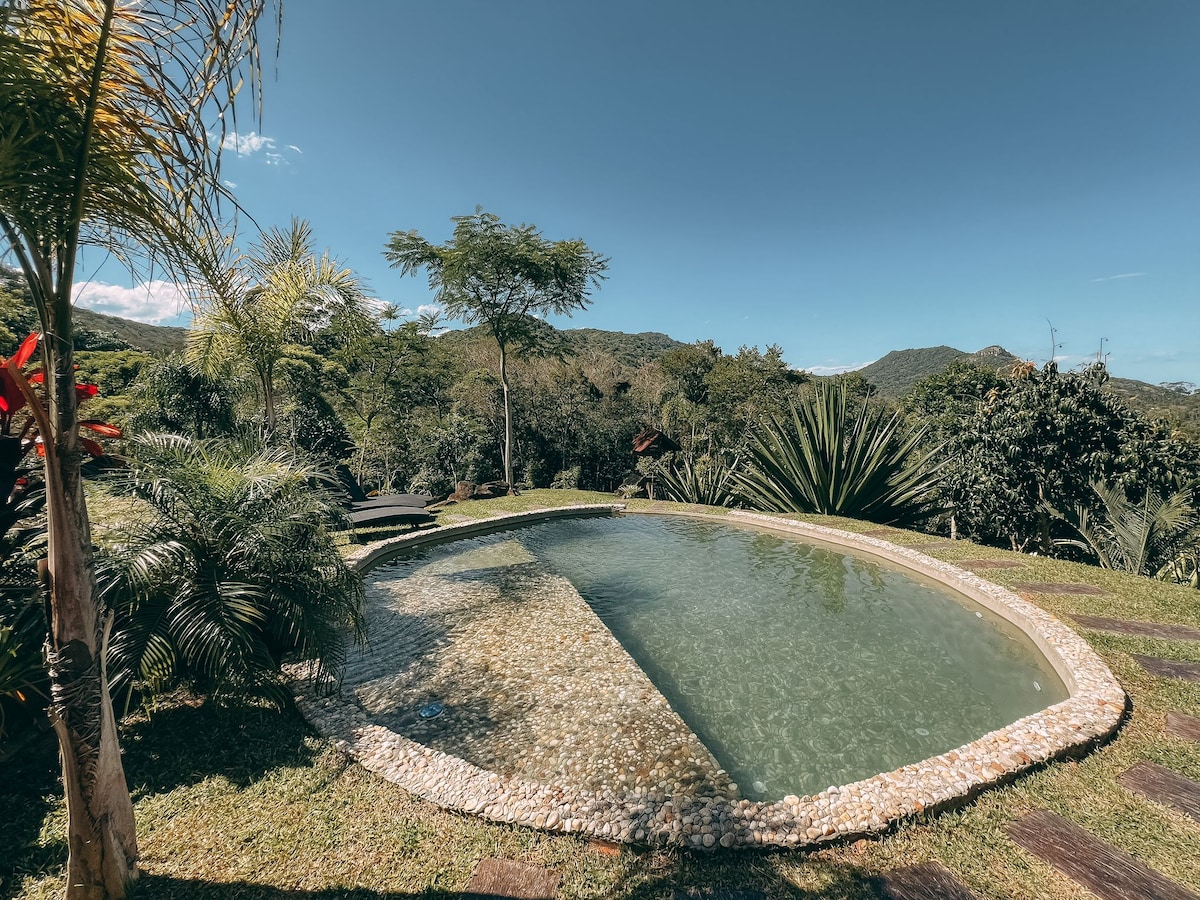 A circular pool is nestled within a landscaped area, surrounded by lush greenery and native plants. The water is calm, reflecting the clear blue sky and distant hills, creating a serene outdoor ambiance. Lounge seating is available nearby for relaxation.