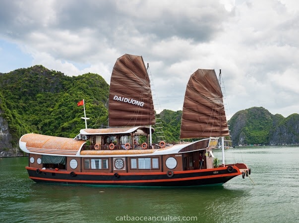 A traditional cruise boat is anchored on calm waters, featuring a distinct dual sail design and wooden features. Lush green mountains rise in the background, creating a serene landscape. The boat showcases multiple outdoor seating areas for guests to relax and enjoy the view.