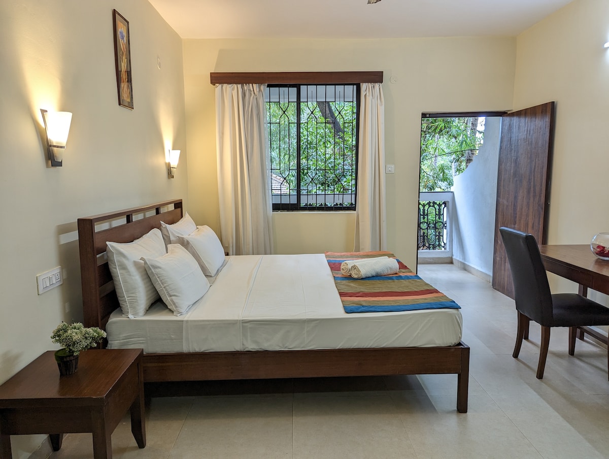 A well-lit bedroom features a spacious bed adorned with neutral-toned linens and a colorful throw. Nearby, a wooden study table and chair are positioned next to a window, allowing natural light. A door leads to a small balcony, enhancing the room's airy ambiance.