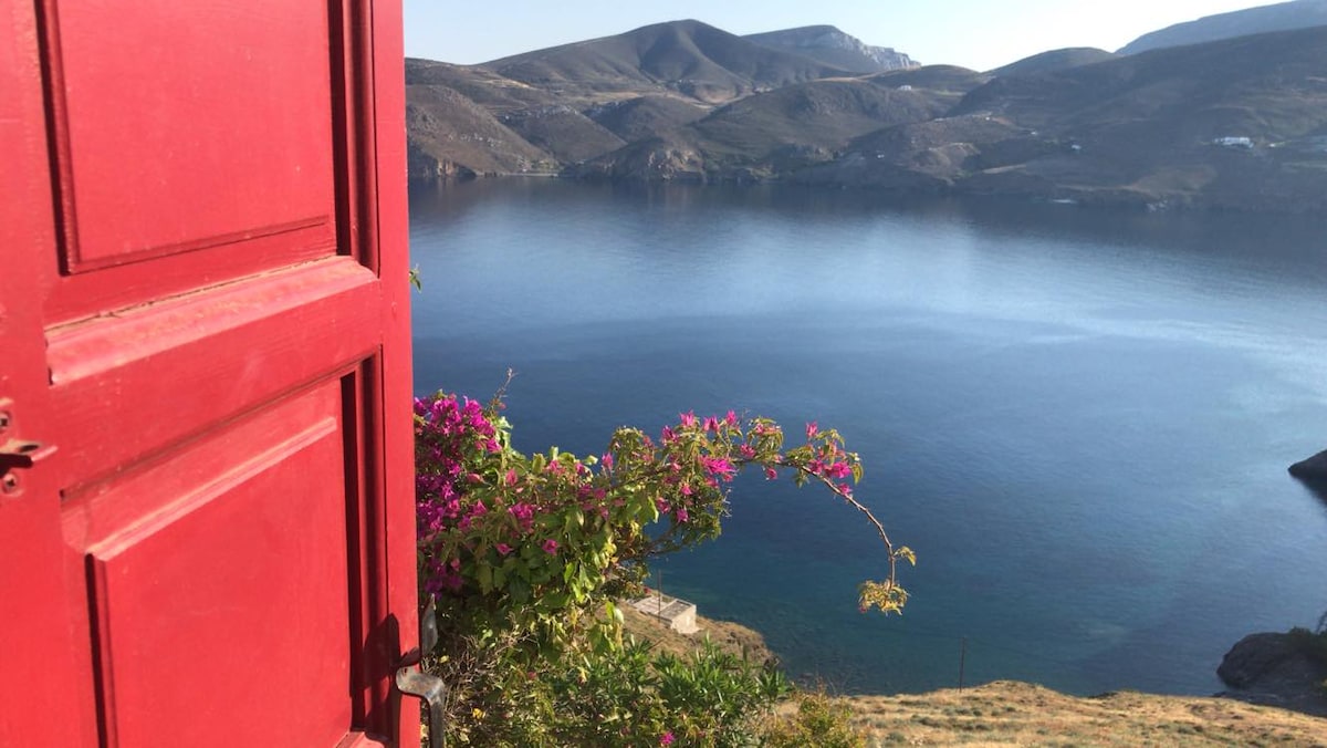A bright red door frames a stunning view of calm waters and distant hills. Bougainvillea flowers add a splash of color, cascading from the doorway. The tranquil Aegean Sea reflects the clear sky, creating a serene setting.