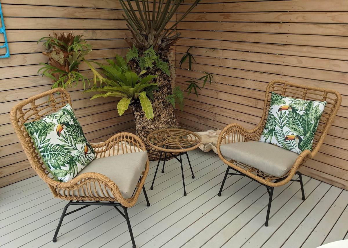A cozy seating area features two rattan chairs adorned with tropical-patterned cushions, positioned around a small round table. Lush greenery is present in the background, creating a serene ambiance. The wooden walls and deck add a natural touch to the space.