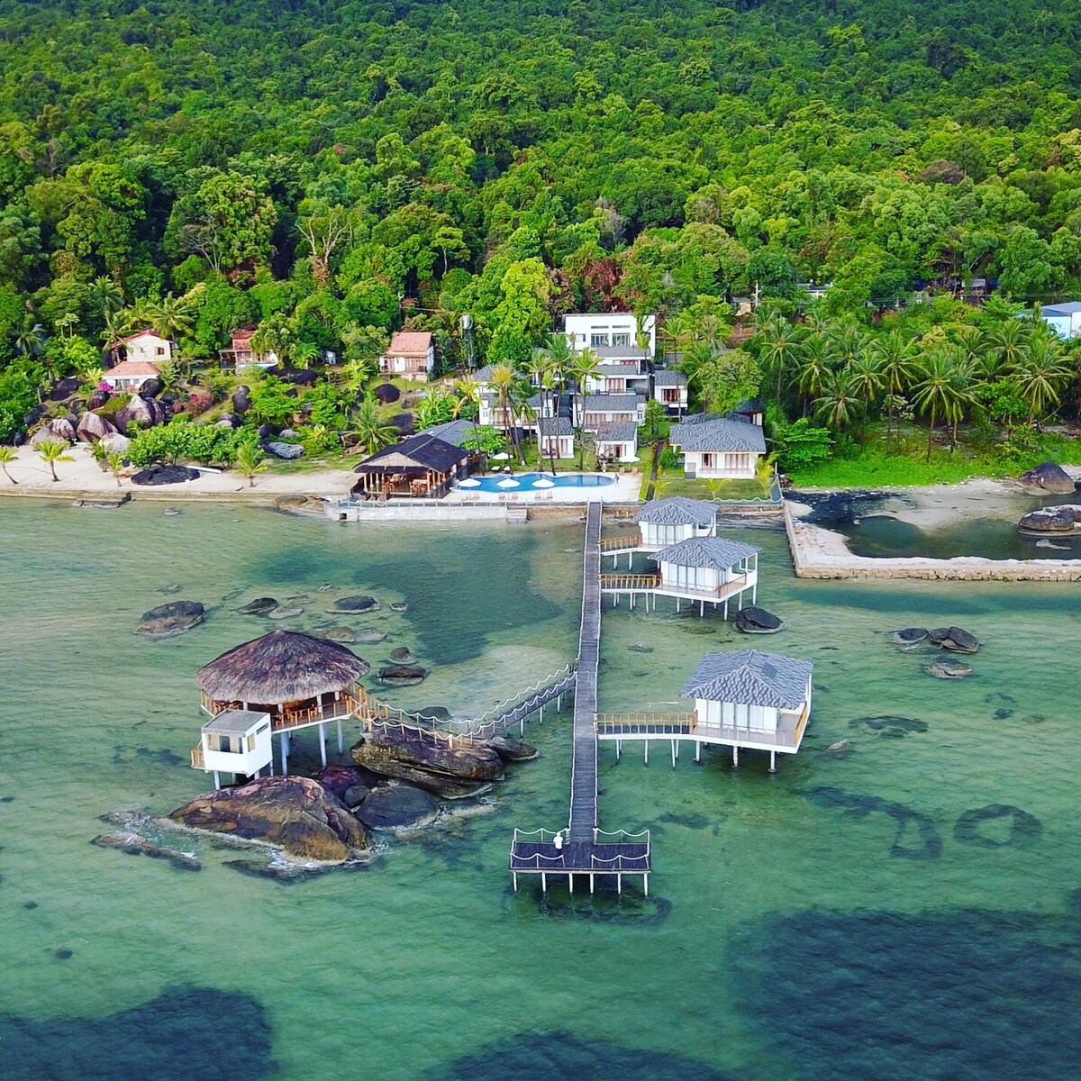An aerial view of The Pier Phu Quoc Resort showcases a wooden pier extending over clear waters, connecting a variety of structures. Lush greenery surrounds the area, with traditional huts and modern buildings positioned along the coastline, enhancing the serene environment.
