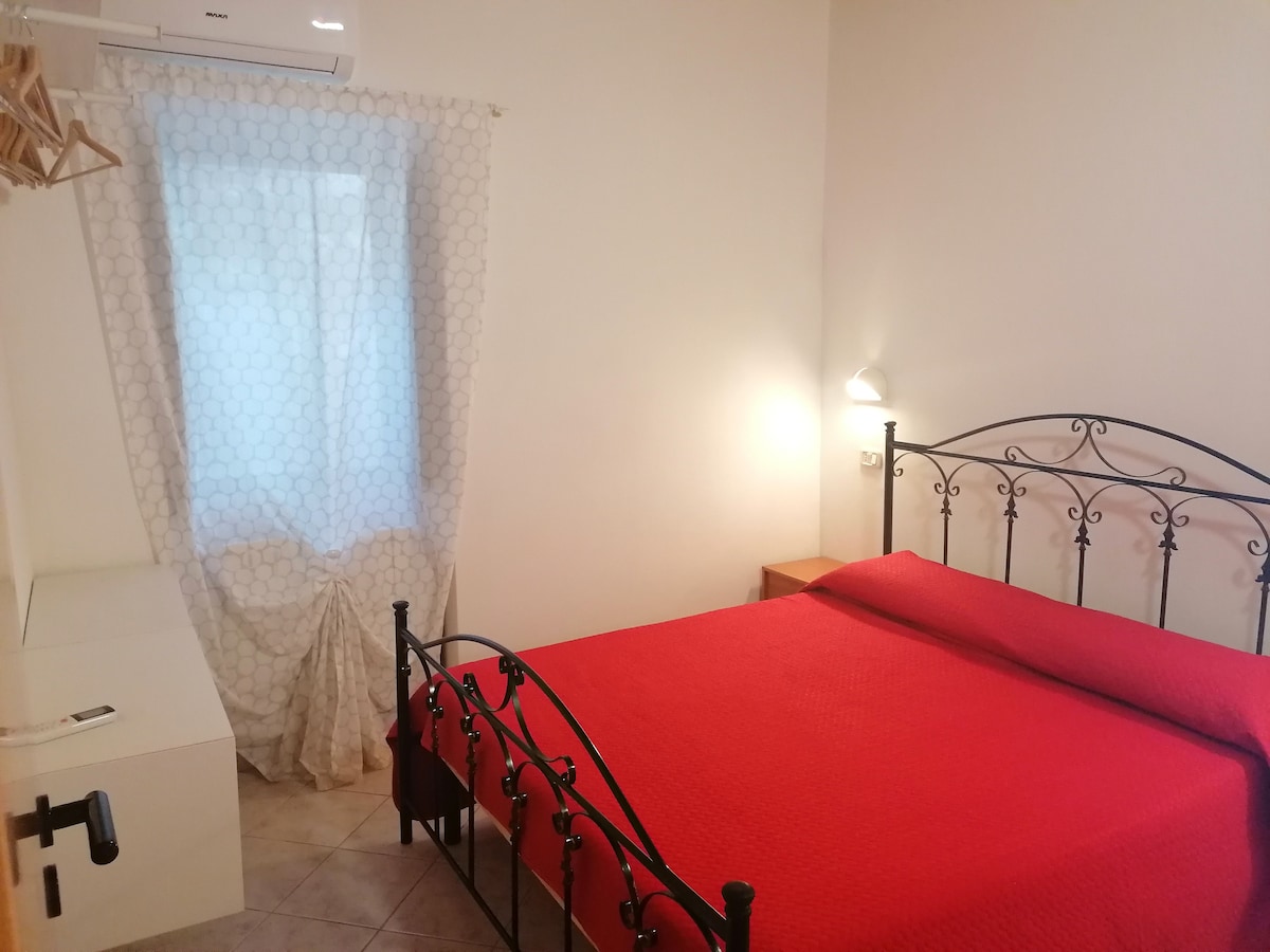A spacious sleeping area features an elegant iron bed with a bright red bedspread. Soft light is provided by a bedside lamp, and a window is dressed in sheer curtains. A small wooden nightstand and a light-colored dresser complete the room's furnishings.