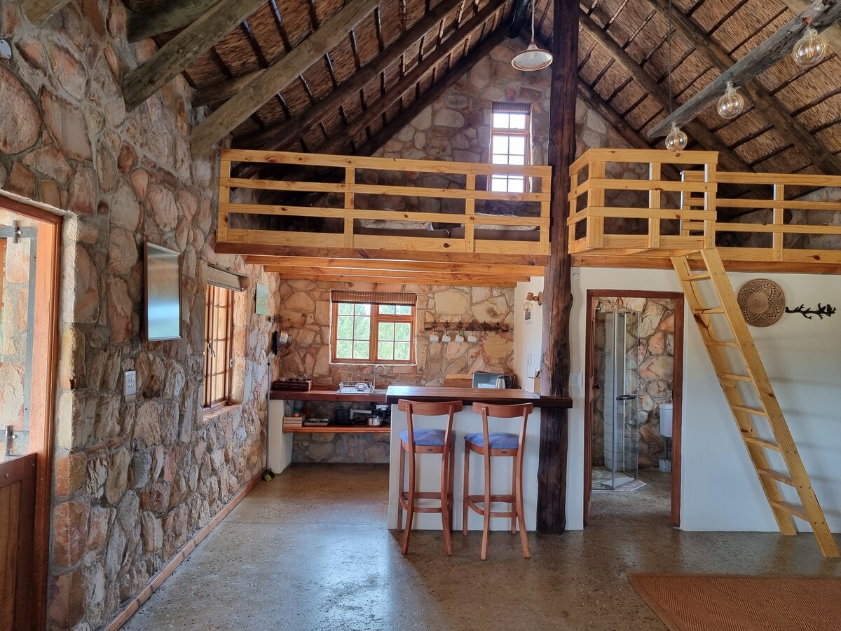 An open-plan living area features exposed stone walls and high ceilings with wooden beams. A well-lit kitchen is equipped with a bar area containing four high stools. A wooden staircase leads to a lofted space, while natural light flows through multiple windows.