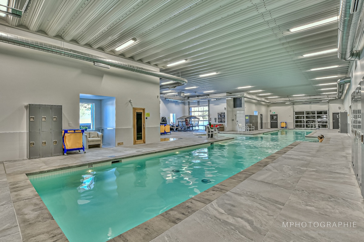 An indoor pool area features a spacious swimming pool with light blue water. The surroundings include changing room lockers and various seating options. Natural light filters in through large windows, illuminating the area, while modern lighting fixtures enhance visibility throughout the space.