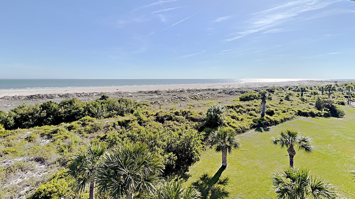 An expansive view captures the coastline, showcasing the sandy beach meeting the ocean under a clear blue sky. Lush greenery and palm trees adorn the foreground, enhancing the natural landscape. The horizon stretches far into the distance, inviting a sense of tranquility.