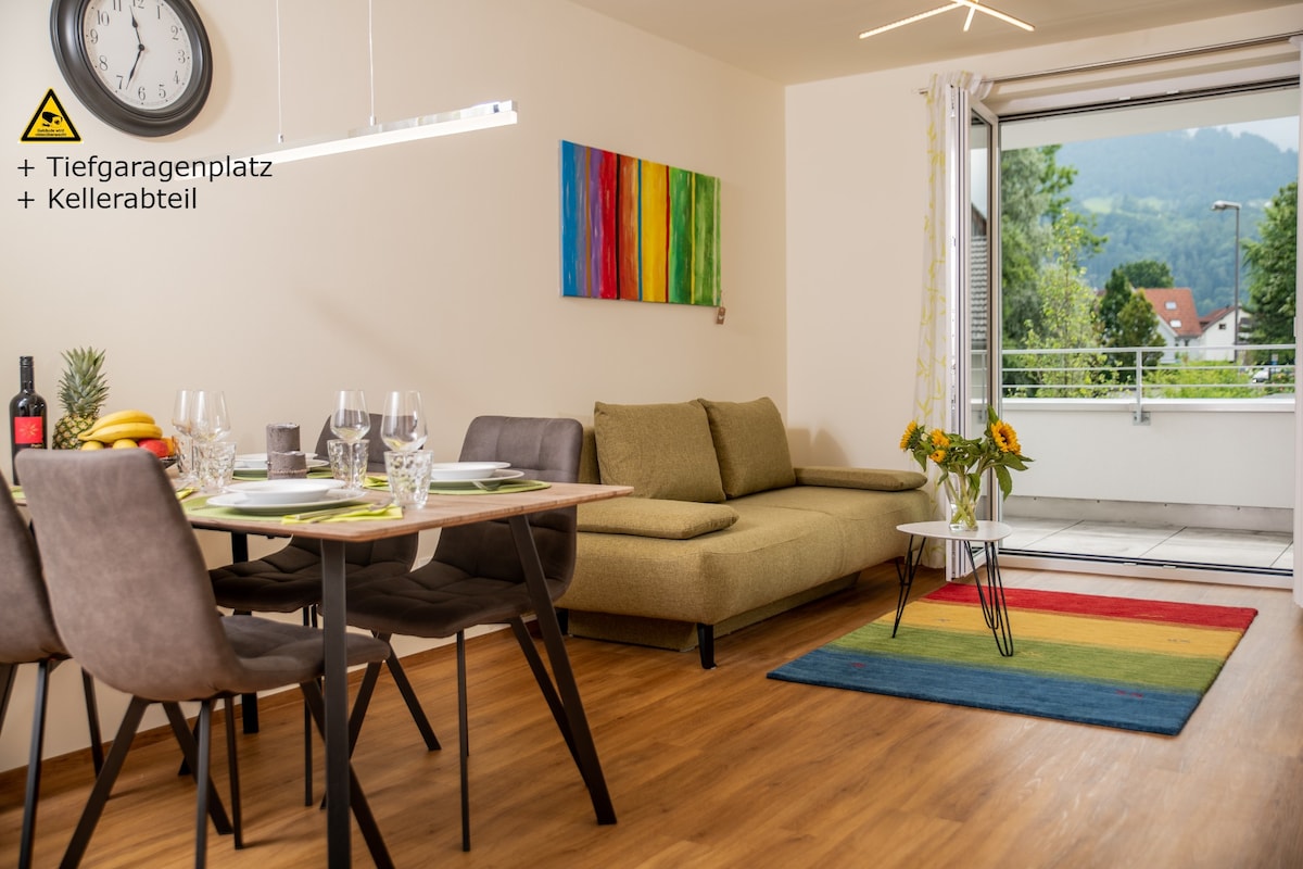 The living and dining area features a comfortable couch and a dining table set for six, with elegant glassware displayed. A sliding door provides access to a balcony with mountain views. Colorful artwork is displayed on the wall, complementing the vibrant area rug.