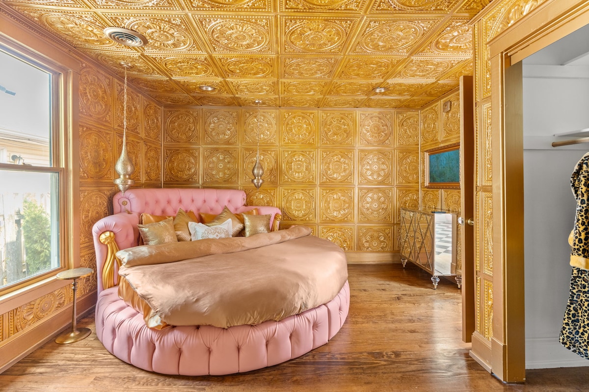 A uniquely designed room showcases a plush round king bed adorned with golden and pink accents. The walls feature intricately patterned gold panels, while light from the windows highlights the warm tones. A television is mounted across from the bed, and elegant lighting fixtures add to the decor.