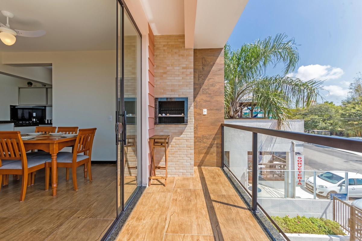 A spacious balcony area is visible, showcasing glass railings and tiled flooring. A barbecue grill is installed in a built-in section. The dining room can be seen through large glass doors, featuring a wooden table and chairs. Bright sunlight illuminates the outdoor space.