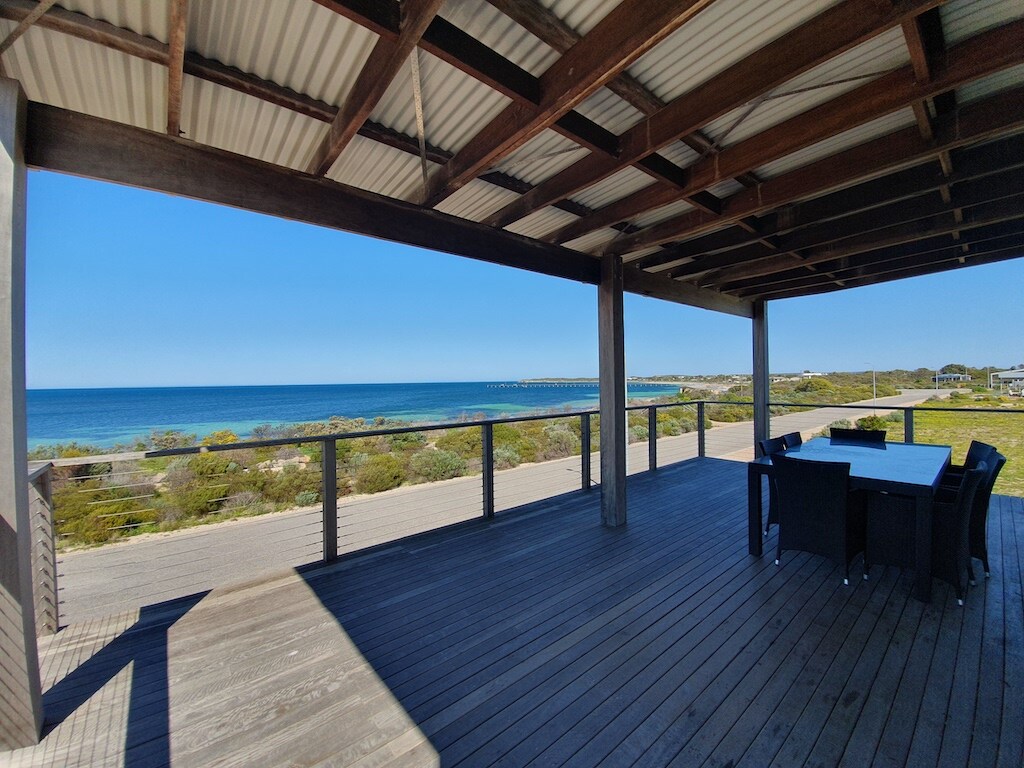 An expansive outdoor deck features a large dining table surrounded by black chairs, overlooking stunning ocean views. An unobstructed horizon showcases calm waters against a clear blue sky, with the coastline visible in the distance. The deck's wooden flooring complements the modern structure.
