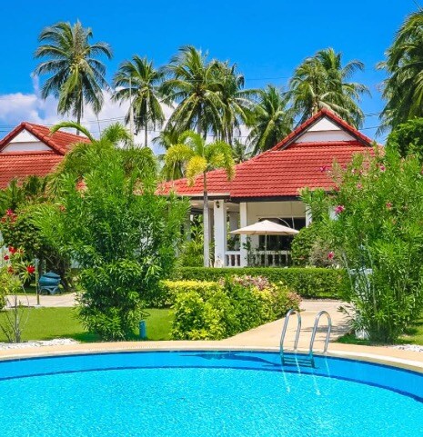 A well-maintained garden surrounds a clear blue pool, with lounge chairs positioned nearby. A house with a red-tiled roof is visible in the background, framed by palm trees and vibrant foliage. An umbrella-covered seating area adds to the inviting outdoor space.