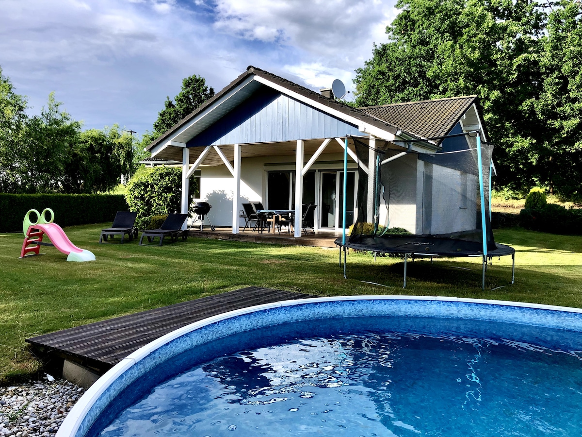 A spacious bungalow is situated amidst lush greenery, featuring a circular swimming pool in the foreground. A trampoline is positioned nearby on a grassy area, alongside a small slide, inviting outdoor play. Large glass doors open to an inviting patio area furnished with seating.