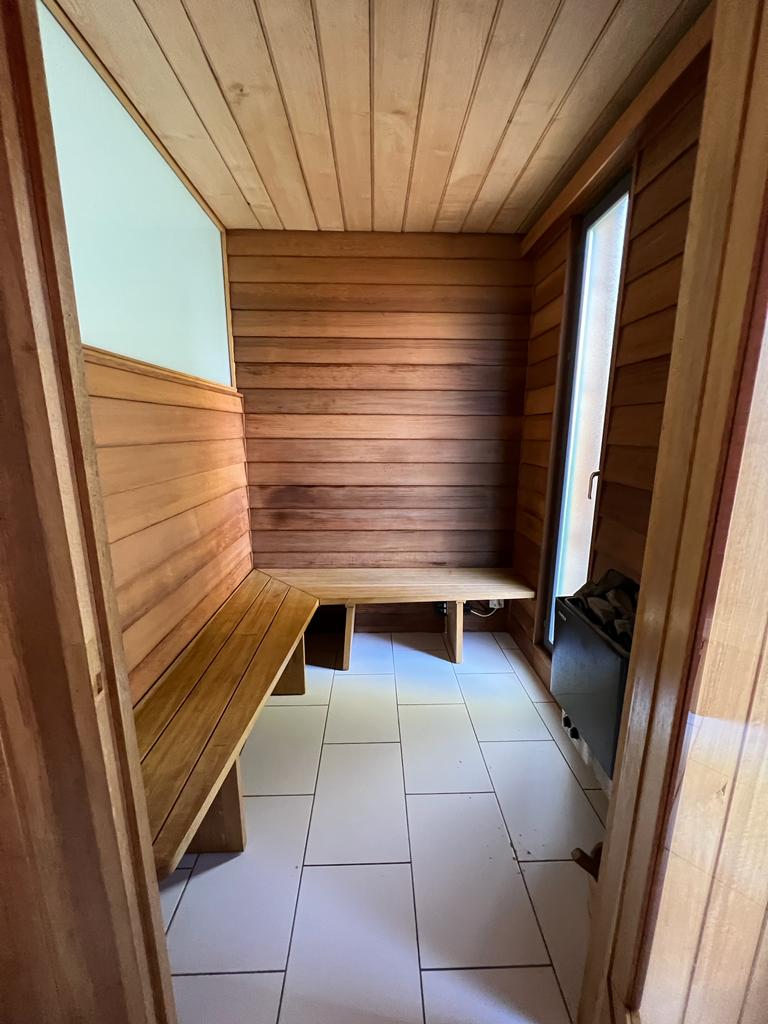 A wooden sauna room is visible, featuring a tiled floor and slatted wooden benches along the walls. Natural light enters through a window, illuminating the warm tones of the wood. The heater is positioned against the far wall, contributing to a serene environment.