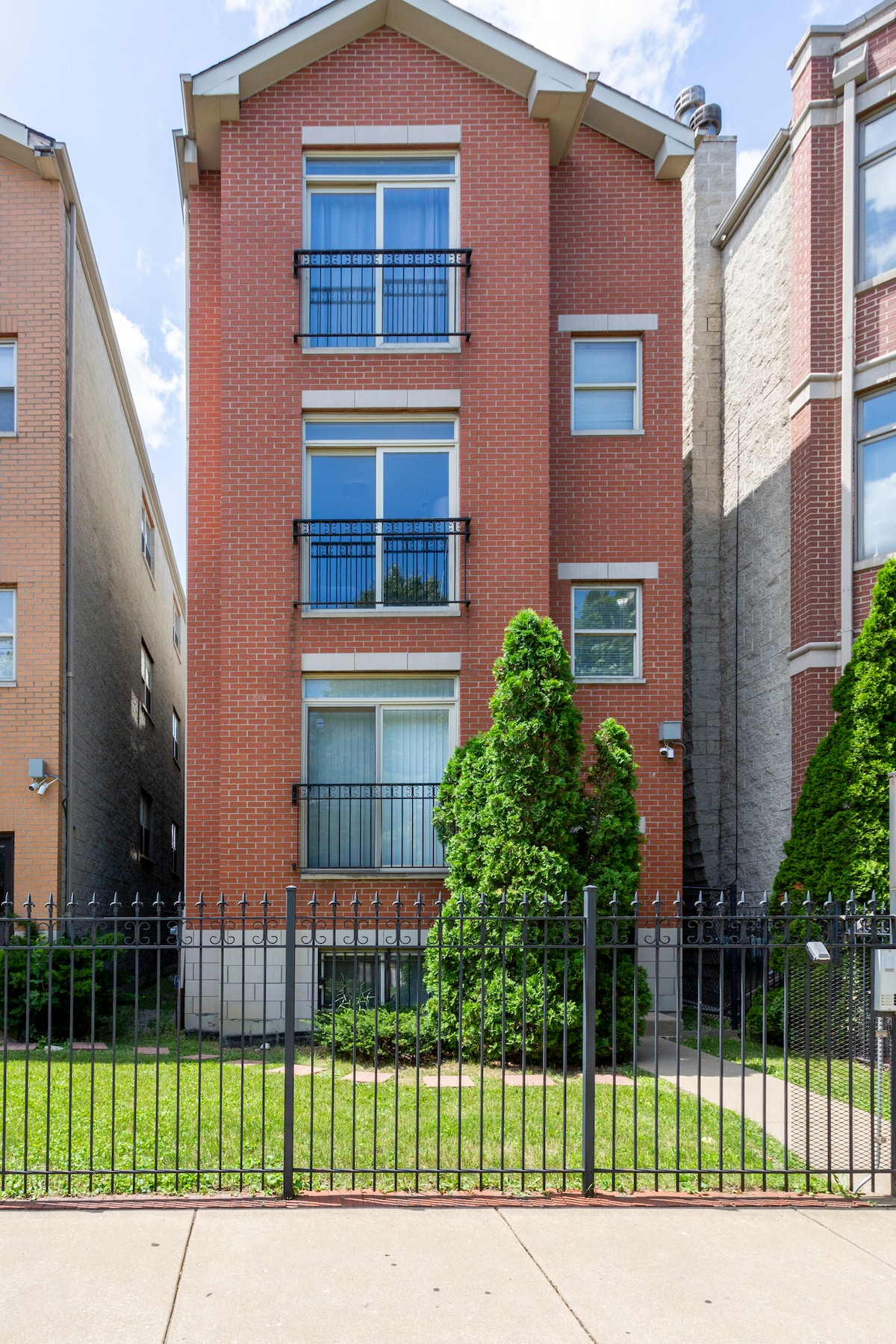 A three-story brick building is surrounded by a well-maintained garden, featuring lush greenery and a manicured lawn. Large windows provide natural light to the interior, while black wrought iron fencing outlines the property for added privacy.