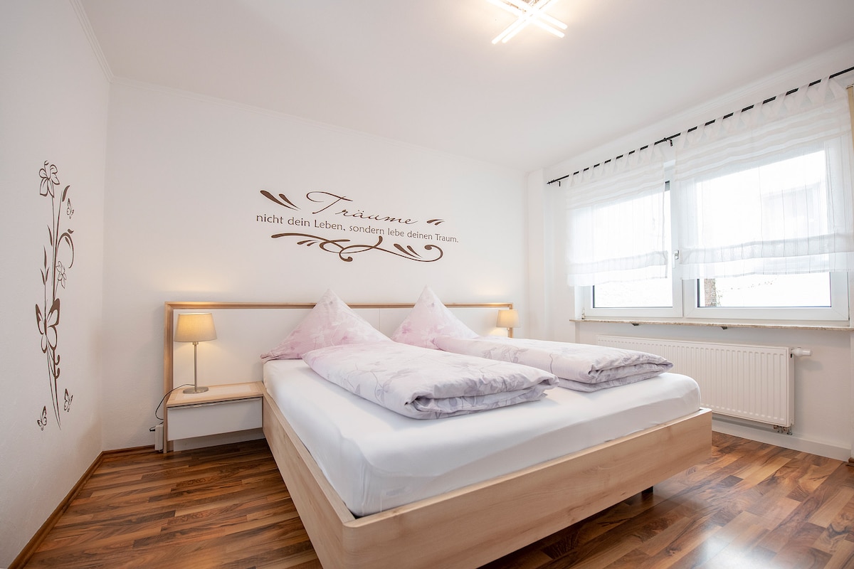 A bright sleeping area features a bed with plush pillows and a light wood frame. Accent lighting beside the bed creates a soft glow. Large windows dressed with sheer curtains allow natural light to fill the space, enhancing the serene environment.