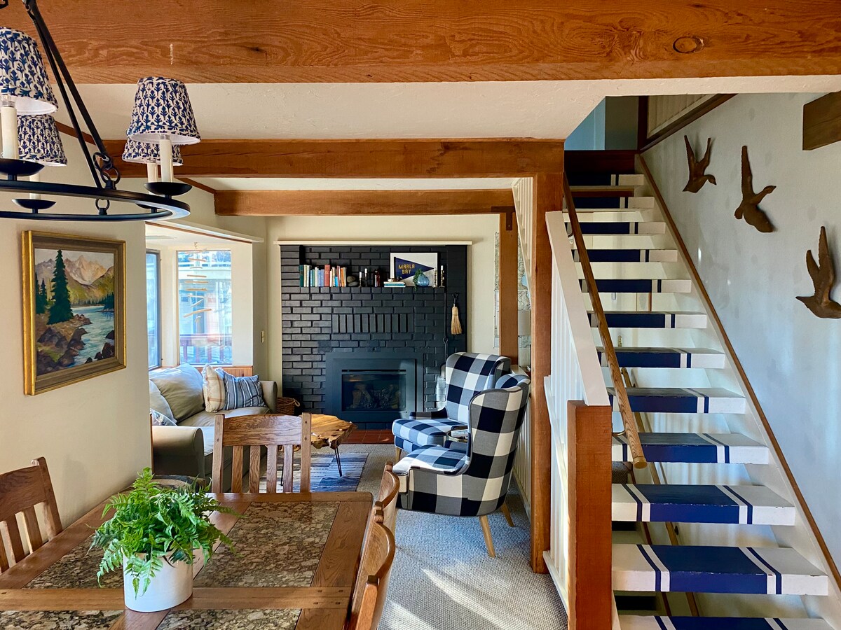 A spacious living area features a cozy arrangement with a black and white checkered chair, a light-colored sofa, and a black brick fireplace. Wooden beams support the ceiling, while a dining table sits beneath a chandelier. A staircase with blue and white accents leads to the upper floors.