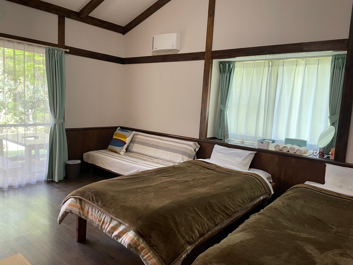 A spacious guest room features two beds with soft brown blankets, alongside a comfortable sofa. Natural light filters through sheer curtains, illuminating the space. A mirror enhances the simplicity, and a doorway leads to a private terrace beyond.