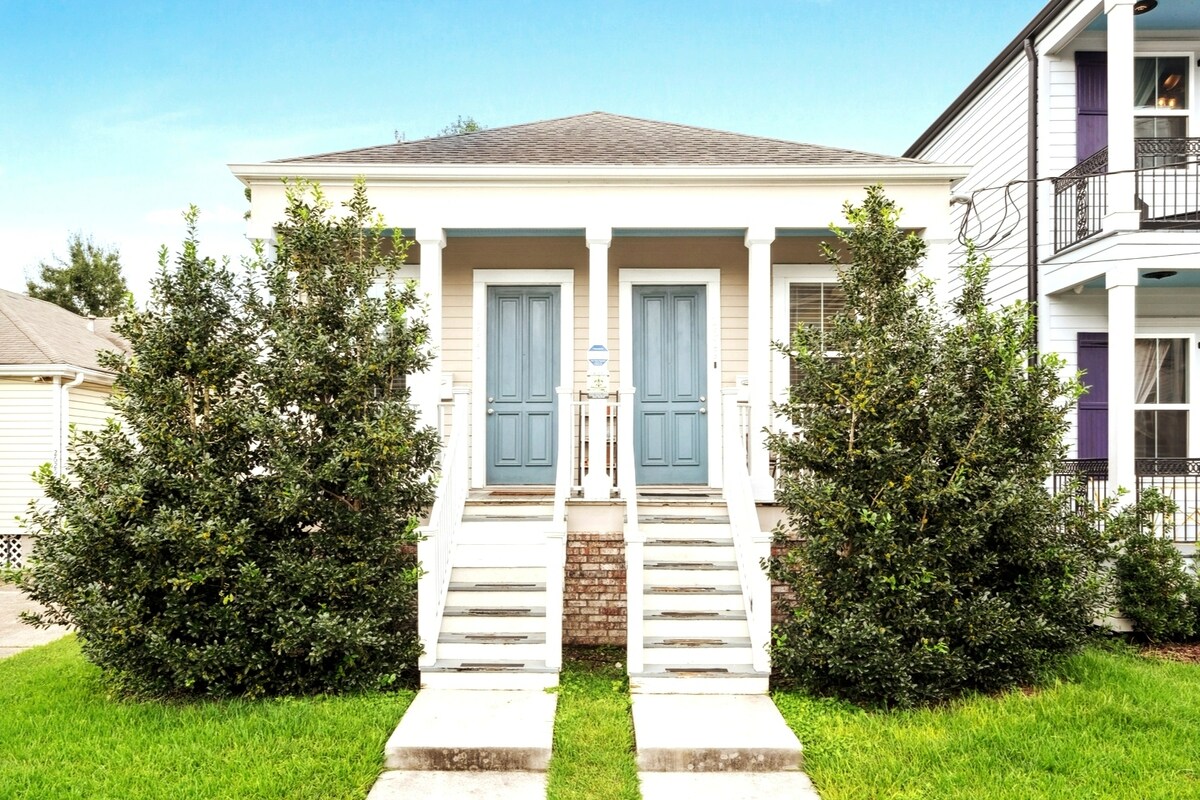 The exterior of a newly-constructed shotgun house is shown, featuring a symmetrical facade with two blue doors. Two sets of concrete steps lead to each entrance, surrounded by neatly trimmed shrubs. A grassy area is visible in the foreground, contributing to a welcoming appearance.