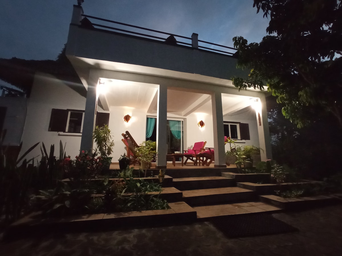 The villa is illuminated at dusk, featuring a welcoming entryway with steps leading to an open porch. Potted plants and tropical greenery frame the space, while outdoor seating is arranged in vibrant colors, creating a cozy atmosphere for relaxation.