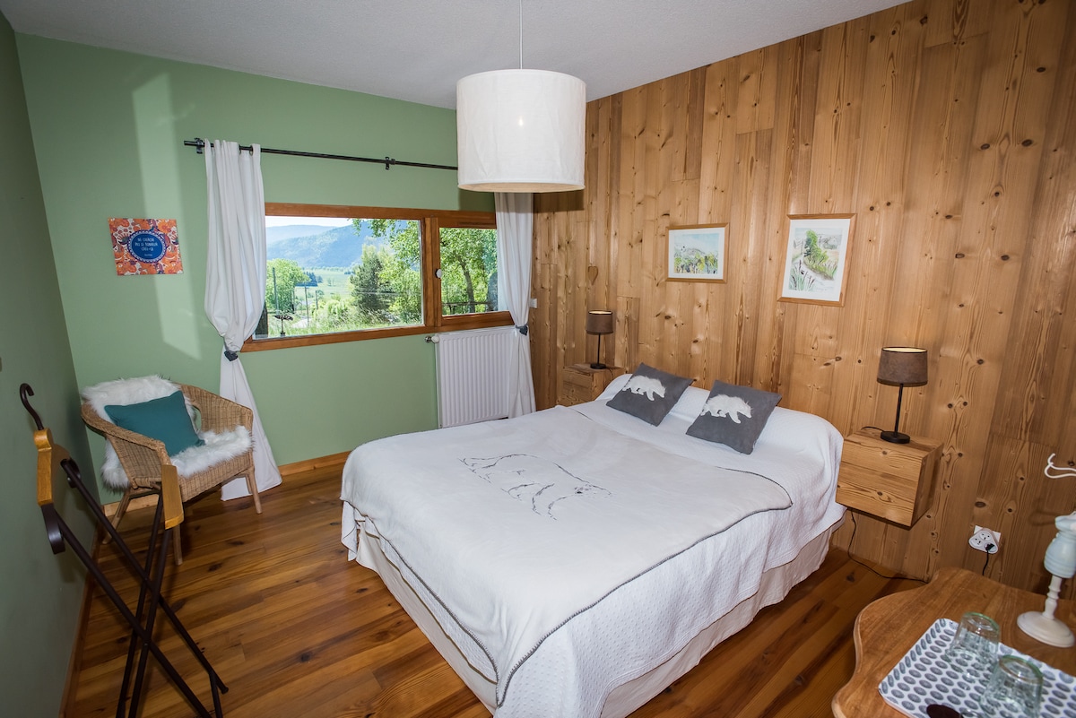 A cozy bedroom features a spacious bed with a light-colored coverlet, complemented by two decorative pillows. Soft green walls and natural wood accents create a warm ambiance. A large window offers views of the surrounding landscape, while natural light brightens the room.