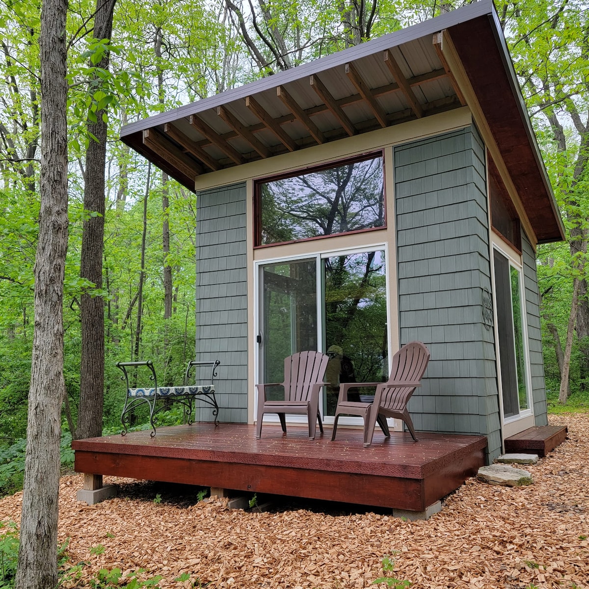 A small cabin is nestled among lush green trees, featuring large windows that allow natural light to illuminate its interior. On the deck, two Adirondack chairs are positioned, inviting guests to relax and enjoy the serene forest surroundings. A gravel walkway leads to the entrance.
