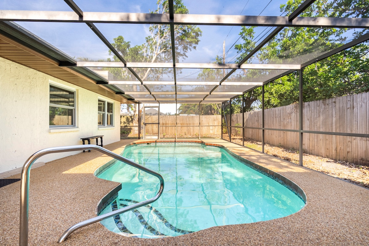 A screened-in pool is featured in the backyard, surrounded by a privacy fence. The pool area has a textured surface and a gently sloped entry, with clear water reflecting the sky. Green foliage and trees are visible in the background, enhancing the serene environment.