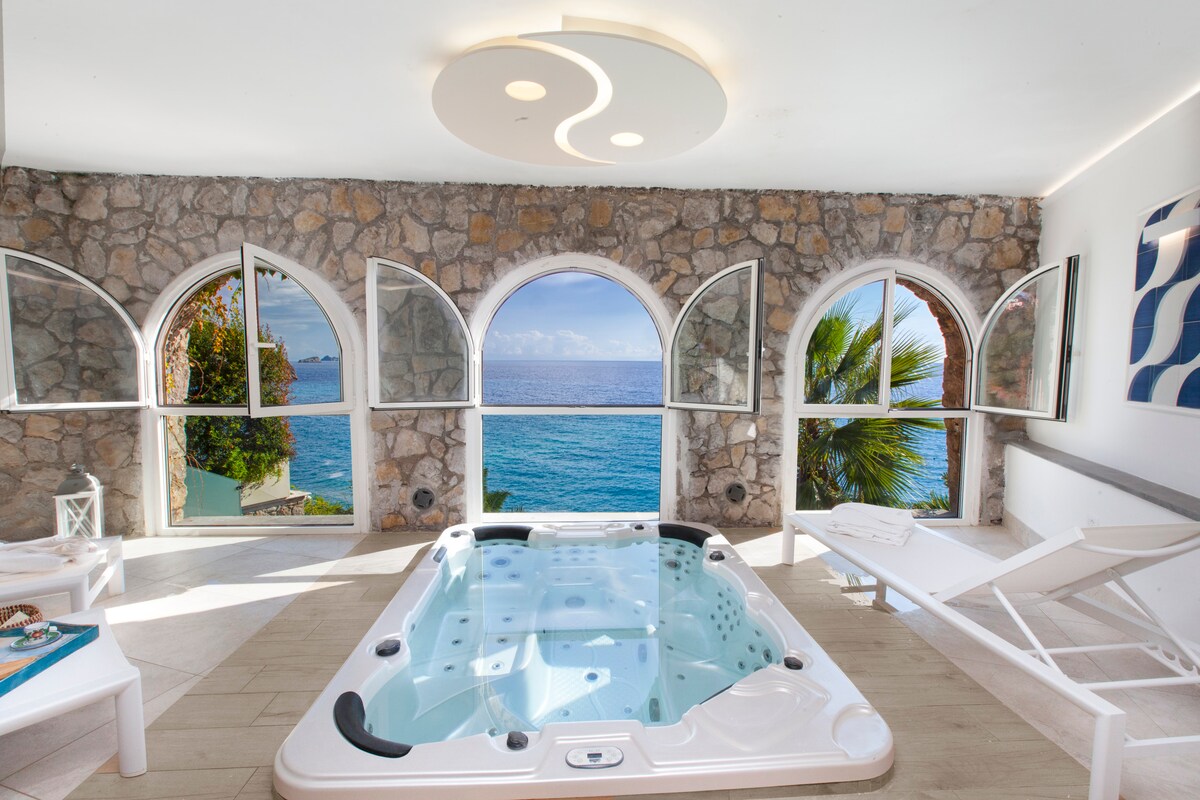 A relaxing indoor spa area is framed by arched windows, which open to reveal a view of the sea. The hot tub is positioned in the center, surrounded by stone walls and minimalist decor. Sunlight floods the space, enhancing the tranquil atmosphere.