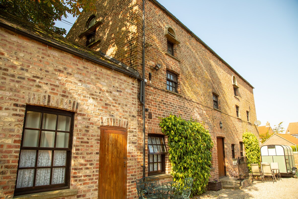 A historic building featuring a blend of brick walls and greenery. The façade showcases large windows, providing ample natural light. A seating area with chairs can be seen in the foreground, surrounded by gravel and a serene outdoor space.