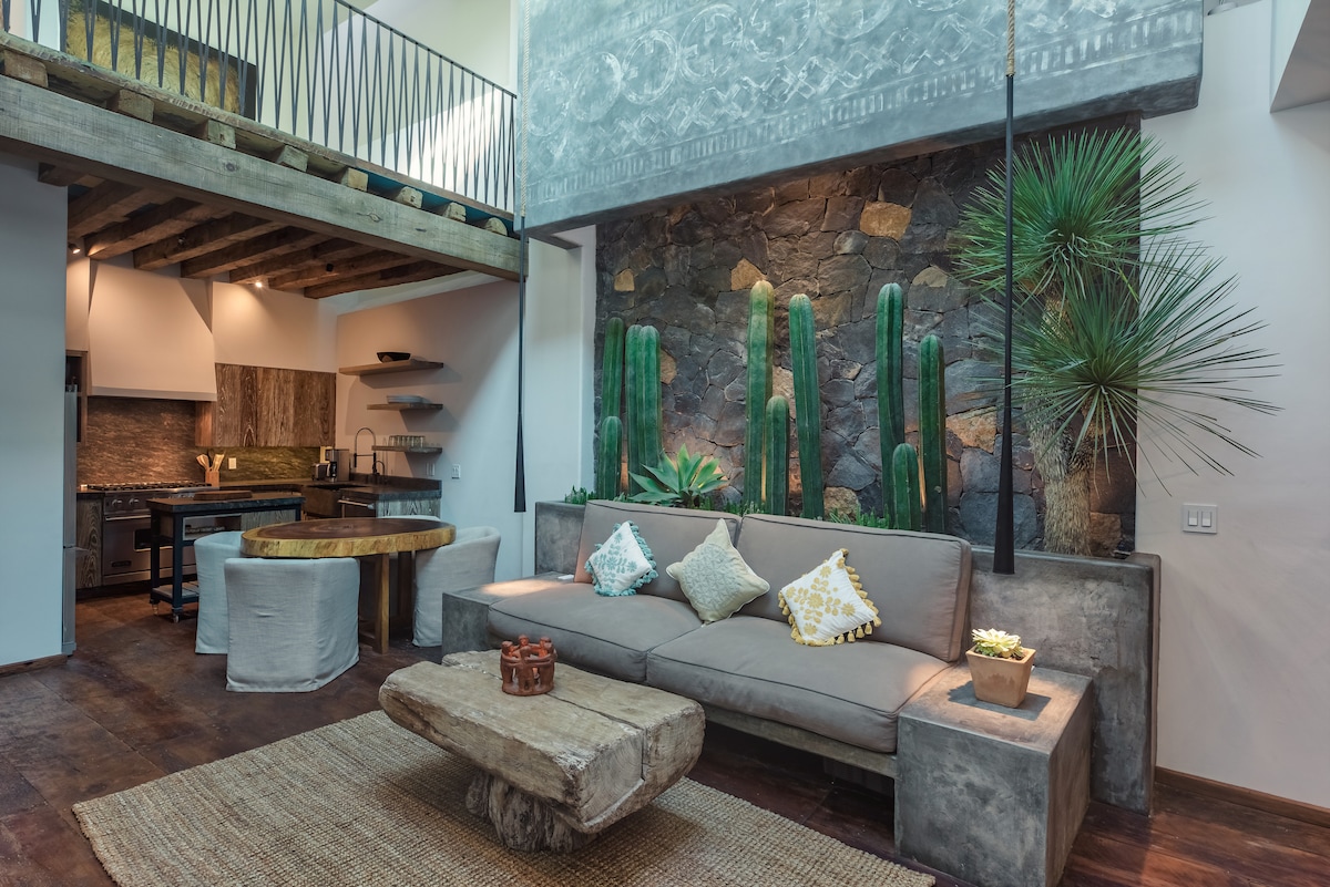 A spacious living area is designed with a comfortable sofa and a natural wood coffee table. A stone wall adorned with cacti adds a touch of greenery, while the kitchen space is visible in the background, featuring modern appliances and rustic finishes.